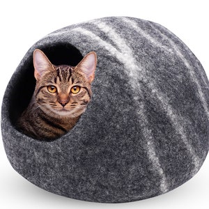 May include: A gray cat bed with a striped pattern, featuring a tabby cat peeking out of the opening. The cat has brown and black stripes and yellow eyes. The bed is a rounded shape, made of a felt-like material.