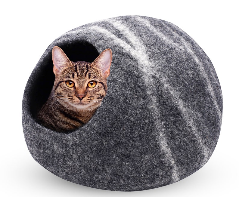May include: A gray cat bed with a striped pattern, featuring a tabby cat peeking out of the opening. The cat has brown and black stripes and yellow eyes. The bed is a rounded shape, made of a felt-like material.