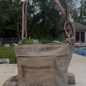 May include: A canvas crossbody bag in shades of olive green and tan, featuring a front pocket with a zipper and the text "VINTAGE 1930." The bag has a brown leather strap and a small patch that says "No. 1."