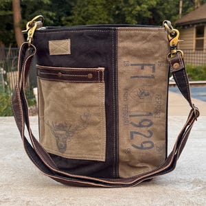 May include: A canvas shoulder bag with a brown leather strap and accents. The bag features a deer head graphic on a tan pocket and the text "F1 1929 ENZO FERRARI" printed on the side. The bag has a black and tan color scheme.
