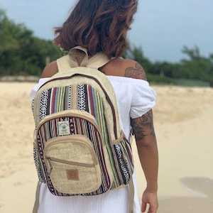 May include: A beige hemp backpack with colorful stripes and a brown leather zipper pull. The backpack has a large main compartment and a smaller front pocket. The brand name "Himalayan Hemp" is printed on the front pocket.