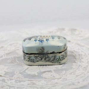 May include: A small, oval-shaped trinket box with a painted scene of a wintery village with blue-domed churches. The box is painted on a white background with black outlines.