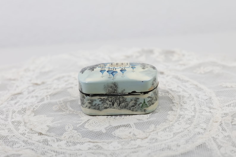 May include: A small, oval-shaped trinket box with a painted scene of a wintery village with blue-domed churches. The box is painted on a white background with black outlines.