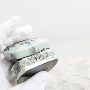 May include: A small, white and blue ceramic box with a winter scene design. The box is open and the lid is resting on top of the box.