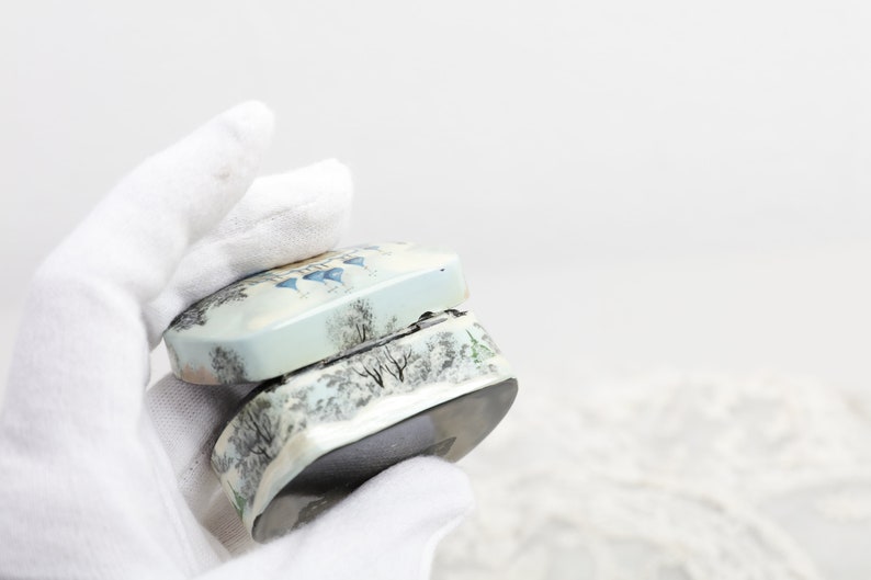 May include: A small, white and blue ceramic box with a winter scene design. The box is open and the lid is resting on top of the box.