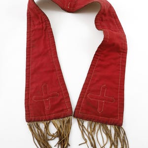 Pode incluir: Uma faixa de tecido vermelho com um desenho de cruz branca bordada em cada extremidade. A faixa tem uma borda com franjas na parte inferior e &eacute; apresentada sobre um fundo branco. Pode ser uma vestimenta religiosa ou um acess&oacute;rio cerimonial.