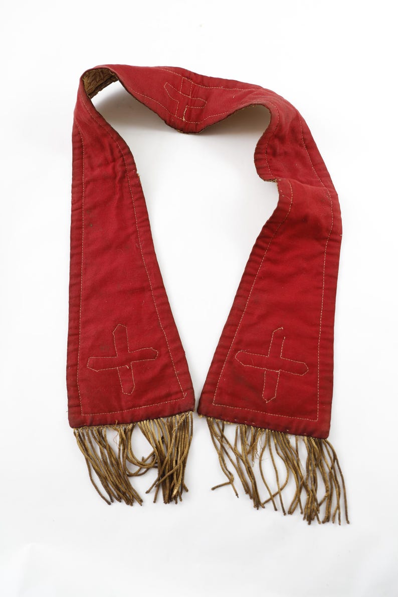 Pode incluir: Uma faixa de tecido vermelho com um desenho de cruz branca bordada em cada extremidade. A faixa tem uma borda com franjas na parte inferior e &eacute; apresentada sobre um fundo branco. Pode ser uma vestimenta religiosa ou um acess&oacute;rio cerimonial.