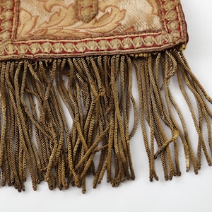 Pode incluir: Close-up de um tecido decorativo com um esquema de cores dourado e vermelho. A parte superior apresenta um padr&atilde;o floral ornamentado, enquanto a parte inferior tem longas franjas douradas torcidas. O tecido faz parte de um item decorativo maior.