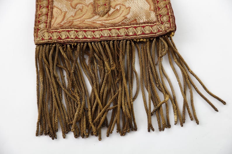Pode incluir: Close-up de um tecido decorativo com um esquema de cores dourado e vermelho. A parte superior apresenta um padr&atilde;o floral ornamentado, enquanto a parte inferior tem longas franjas douradas torcidas. O tecido faz parte de um item decorativo maior.