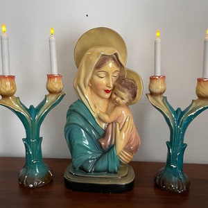 Peut inclure: Une statue en c&eacute;ramique vintage de la Vierge Marie tenant l'enfant J&eacute;sus, flanqu&eacute;e de deux bougeoirs avec des bougies allum&eacute;es. Les bougeoirs sont en c&eacute;ramique et pr&eacute;sentent un motif floral vert et jaune.