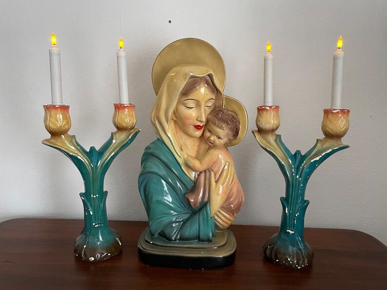 Peut inclure: Une statue en c&eacute;ramique vintage de la Vierge Marie tenant l'enfant J&eacute;sus, flanqu&eacute;e de deux bougeoirs avec des bougies allum&eacute;es. Les bougeoirs sont en c&eacute;ramique et pr&eacute;sentent un motif floral vert et jaune.