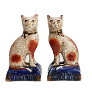 May include: Two ceramic cat figurines with a cream-colored crackle glaze finish, featuring orange-red patches on their bodies and necks. Each cat wears a dark collar with a bow and sits upright on a rectangular, deep blue base with gold trim.