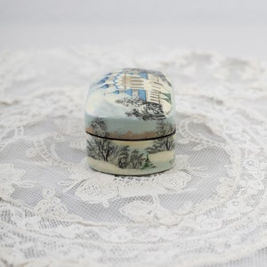 May include: A small, oval-shaped wooden box with a painted scene of a building and trees in a snowy landscape. The box is painted in shades of blue, white, and green.