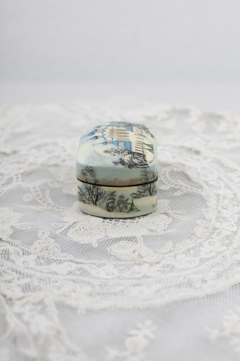 May include: A small, oval-shaped wooden box with a painted scene of a building and trees in a snowy landscape. The box is painted in shades of blue, white, and green.