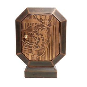 Puede incluir: Un gabinete metálico octogonal de tono bronce con una puerta abatible que presenta un relieve de una flor grande y líneas verticales. El gabinete tiene un pequeño pomo metálico redondo en el lado izquierdo de la puerta y se asienta sobre una base rectangular.