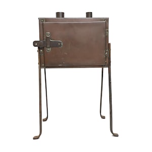 May include: A rectangular brown metal camping stove with a front-loading door, a horizontal latch mechanism, and four thin, outward-curving metal legs. Two cylindrical exhaust ports are visible on the top surface of the stove body.