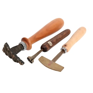 Op de afbeelding: Drie antieke metalen gereedschappen met houten handvatten. De gereedschappen omvatten een stempel met een bloemenontwerp, een gereedschap met een afgerond uiteinde en een gereedschap met een plat, gebogen uiteinde. De handvatten zijn lichtbruin en donkerbruin.