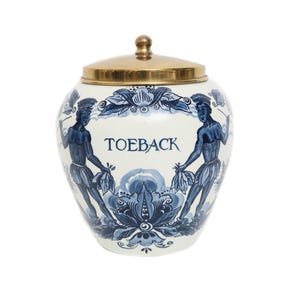 Pode incluir: Jarro de cerâmica branco e azul com tampa dourada. O jarro apresenta um desenho floral azul e duas figuras segurando folhas. A palavra "TOEBACK" está escrita em azul no jarro.
