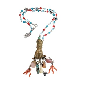 Puede incluir: Un collar con una cadena de cuentas rojas y azul claro alternadas. El colgante es una mano de metal color bronce que sujeta un grupo de dijes, incluyendo dos ramas color coral, un zapato pequeño y varias piedras lisas y pulidas.