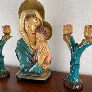 Peut inclure: Une statue vintage de la Vierge Marie tenant l'enfant J&eacute;sus, avec une aur&eacute;ole au-dessus de sa t&ecirc;te. La statue est peinte dans des tons de bleu, blanc et or. Deux bougeoirs assortis avec des sommets en forme de tulipe sont de chaque c&ocirc;t&eacute; de la statue.