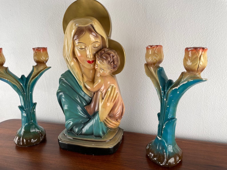 Peut inclure: Une statue vintage de la Vierge Marie tenant l'enfant J&eacute;sus, avec une aur&eacute;ole au-dessus de sa t&ecirc;te. La statue est peinte dans des tons de bleu, blanc et or. Deux bougeoirs assortis avec des sommets en forme de tulipe sont de chaque c&ocirc;t&eacute; de la statue.
