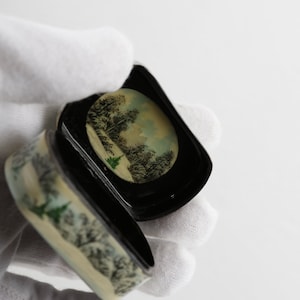 May include: A small, round, black and white trinket box with a winter scene painted on the lid. The box is open and the inside is black.