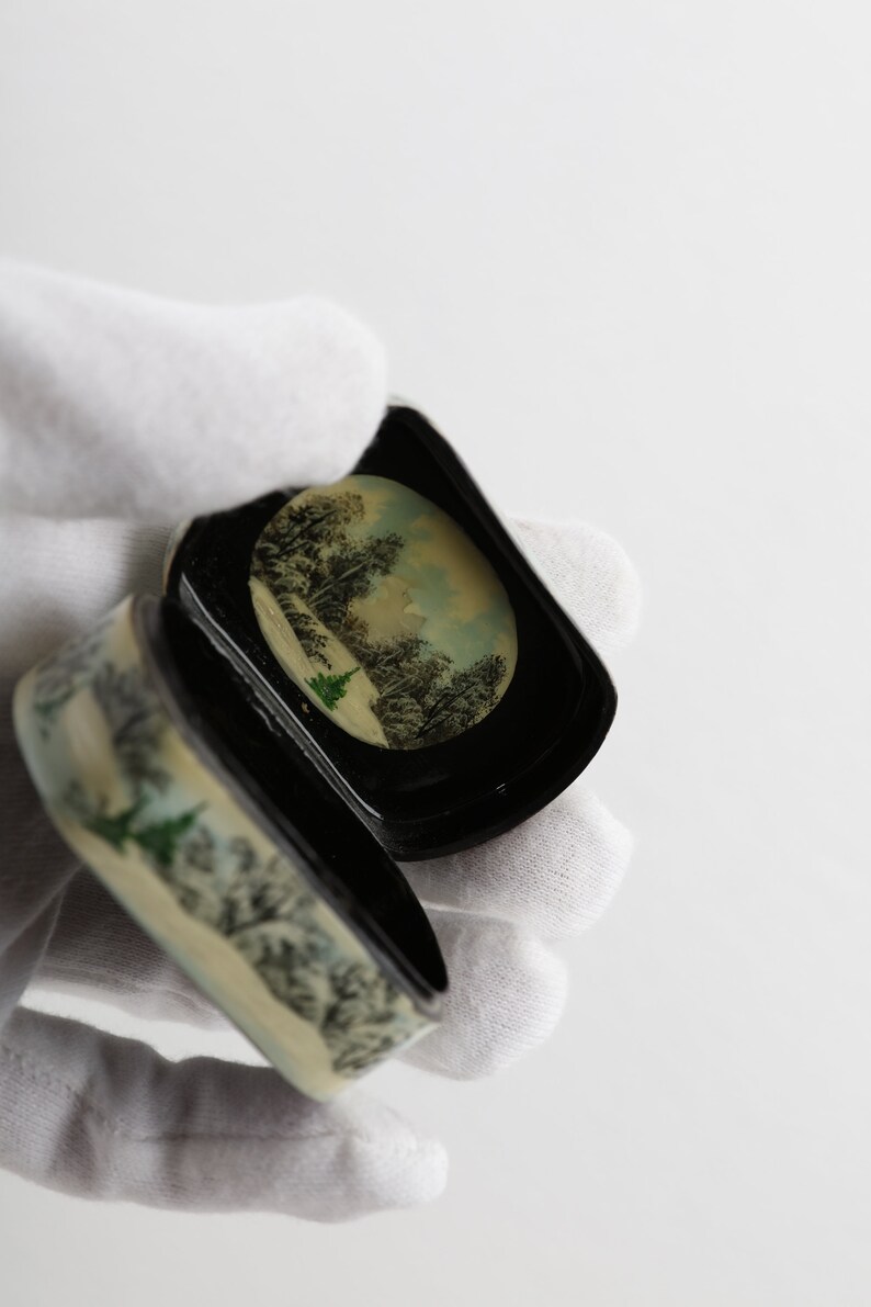 May include: A small, round, black and white trinket box with a winter scene painted on the lid. The box is open and the inside is black.
