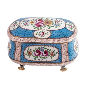 May include: A decorative, oval-shaped trinket box with a light blue background and floral designs. The box features a dotted border and is supported by three small, gold-colored feet. The lid and sides are adorned with oval panels of flowers.