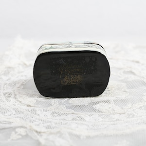 May include: A silver and black rectangular box with a decorative design on the top and bottom. The box is sitting on a white lace surface.