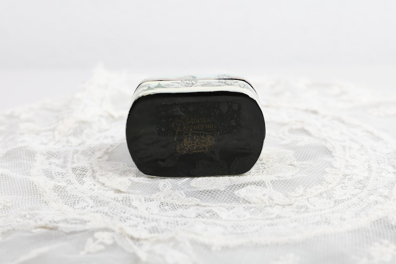 May include: A silver and black rectangular box with a decorative design on the top and bottom. The box is sitting on a white lace surface.