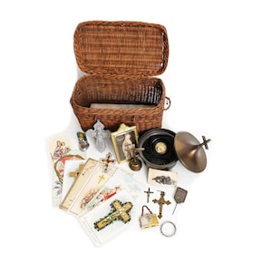 Peut inclure: Un panier en osier tressé marron avec un couvercle ouvert, accompagné d'une collection d'objets religieux comprenant plusieurs cartes de prière en papier illustrées, un petit crucifix en métal, un portrait encadré, un récipient rond et une clochette.