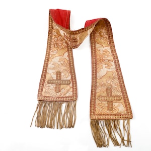 Pode incluir: Uma estola religiosa com gola vermelha e corpo bege. A estola apresenta um desenho de cruz, padr&otilde;es florais e franjas decorativas. As bordas s&atilde;o adornadas com fio vermelho e dourado.