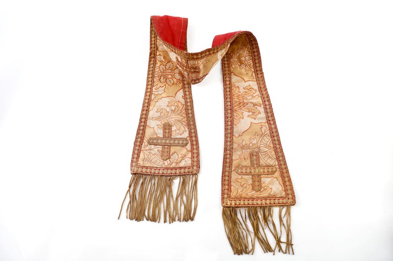 Pode incluir: Uma estola religiosa com gola vermelha e corpo bege. A estola apresenta um desenho de cruz, padr&otilde;es florais e franjas decorativas. As bordas s&atilde;o adornadas com fio vermelho e dourado.