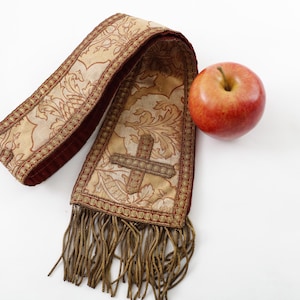 Pode incluir: Uma estola religiosa ornamentada, bege e bord&ocirc;, com um desenho de cruz e franjas, ao lado de uma ma&ccedil;&atilde; vermelha. A estola apresenta padr&otilde;es florais detalhados e &eacute; provavelmente usada para fins cerimoniais.