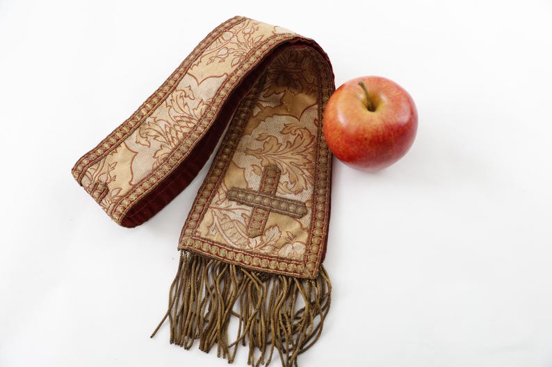 Pode incluir: Uma estola religiosa ornamentada, bege e bord&ocirc;, com um desenho de cruz e franjas, ao lado de uma ma&ccedil;&atilde; vermelha. A estola apresenta padr&otilde;es florais detalhados e &eacute; provavelmente usada para fins cerimoniais.