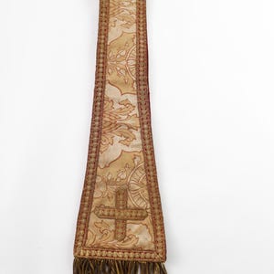 Pode incluir: Uma estola religiosa dourada e bord&ocirc; com um desenho de cruz. A estola apresenta um padr&atilde;o floral ornamentado e uma franja decorativa na parte inferior. O tecido tem uma apar&ecirc;ncia texturizada.