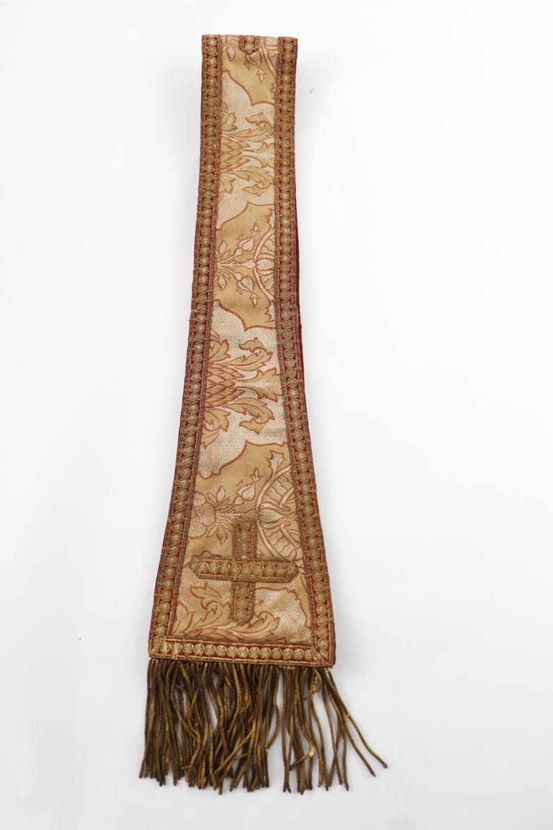 Pode incluir: Uma estola religiosa dourada e bord&ocirc; com um desenho de cruz. A estola apresenta um padr&atilde;o floral ornamentado e uma franja decorativa na parte inferior. O tecido tem uma apar&ecirc;ncia texturizada.