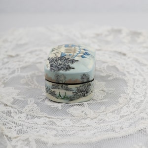 May include: A small, oval-shaped trinket box with a painted scene of a wintery cityscape. The box is decorated with a light blue, white, and gray color scheme. The lid of the box is closed.