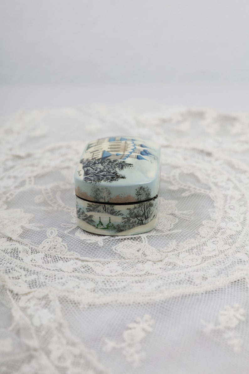 May include: A small, oval-shaped trinket box with a painted scene of a wintery cityscape. The box is decorated with a light blue, white, and gray color scheme. The lid of the box is closed.
