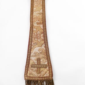 Pode incluir: Uma estola religiosa com um padr&atilde;o floral dourado e creme, uma borda vermelha e um desenho de cruz. A estola tem franjas na parte inferior. O fundo &eacute; branco.