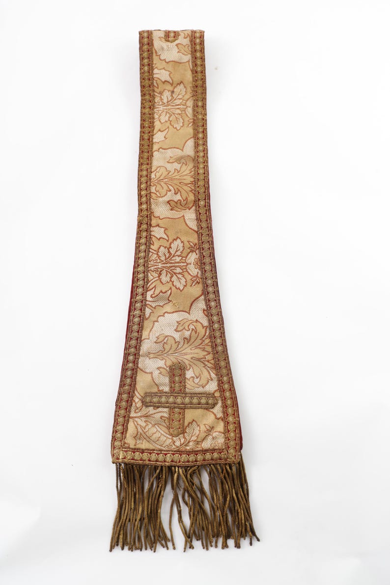 Pode incluir: Uma estola religiosa com um padr&atilde;o floral dourado e creme, uma borda vermelha e um desenho de cruz. A estola tem franjas na parte inferior. O fundo &eacute; branco.