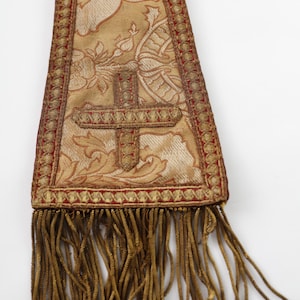 Pode incluir: Uma estola religiosa dourada e vermelha com um desenho de cruz. A estola apresenta intrincados padr&otilde;es florais e uma franja decorativa. O tecido tem uma apar&ecirc;ncia texturizada e a cruz &eacute; bordada.