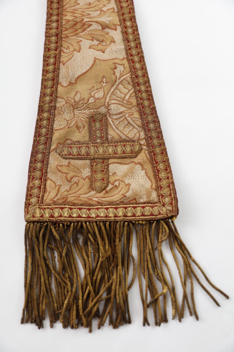 Pode incluir: Uma estola religiosa dourada e vermelha com um desenho de cruz. A estola apresenta intrincados padr&otilde;es florais e uma franja decorativa. O tecido tem uma apar&ecirc;ncia texturizada e a cruz &eacute; bordada.
