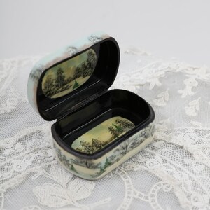 May include: A small, oval-shaped wooden box with a black interior. The box is painted with a winter scene of trees and snow. The lid is open, revealing the empty interior.