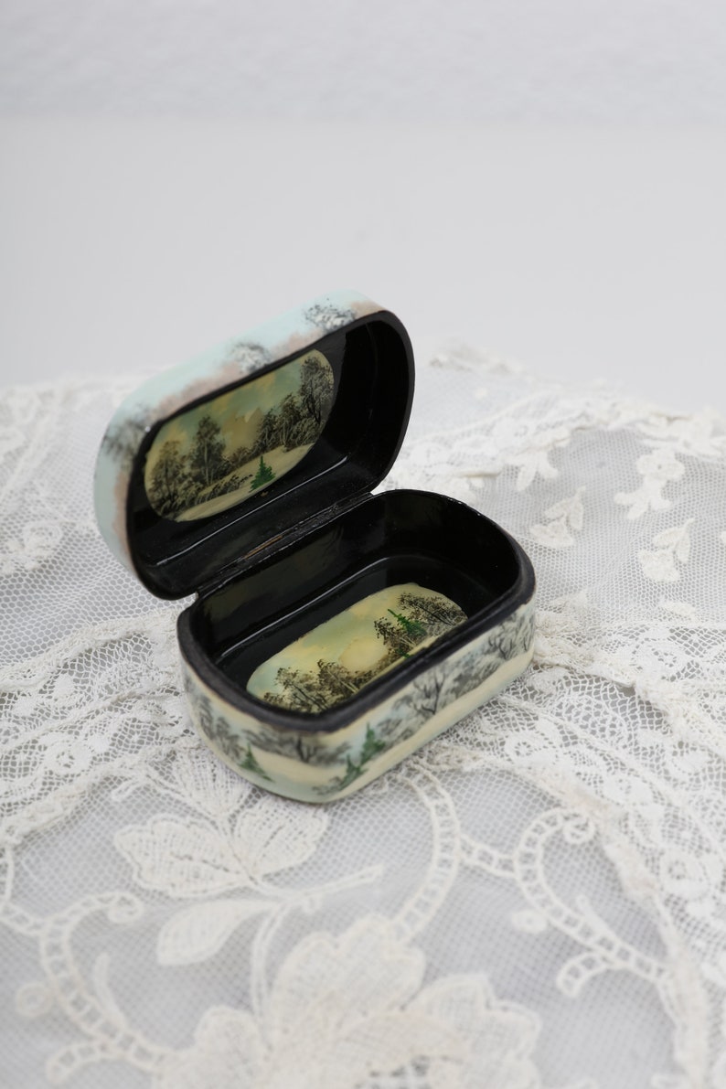 May include: A small, oval-shaped wooden box with a black interior. The box is painted with a winter scene of trees and snow. The lid is open, revealing the empty interior.