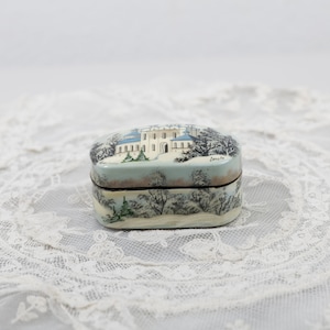 May include: A small, oval-shaped wooden box with a painted scene of a white building with blue accents in a snowy winter landscape. The box has a dark brown lid and a light brown base.