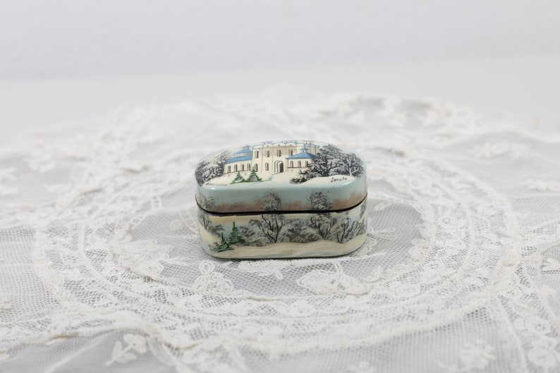May include: A small, oval-shaped wooden box with a painted scene of a white building with blue accents in a snowy winter landscape. The box has a dark brown lid and a light brown base.