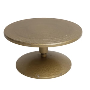 Può includere: Un espositore rotante in metallo color oro dotato di una piattaforma superiore circolare e di una base arrotondata e robusta. La superficie del supporto presenta una finitura opaca testurizzata con cerchi concentrici visibili sul piatto.