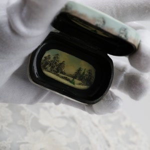 May include: A small, oval-shaped, black and white box with a hinged lid. The lid is open, revealing a painted scene of a winter forest with trees and a stream. The box is held in a white gloved hand.