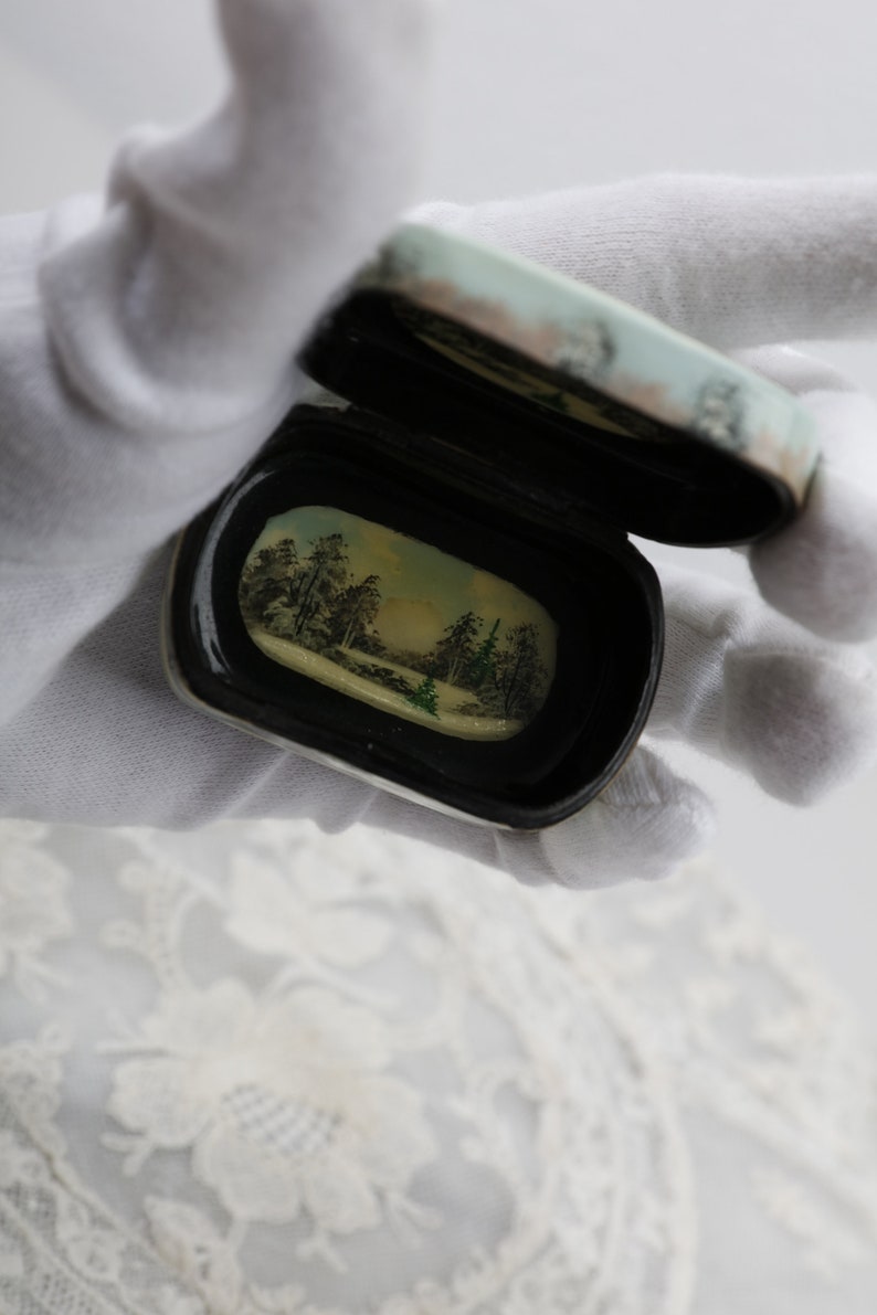 May include: A small, oval-shaped, black and white box with a hinged lid. The lid is open, revealing a painted scene of a winter forest with trees and a stream. The box is held in a white gloved hand.